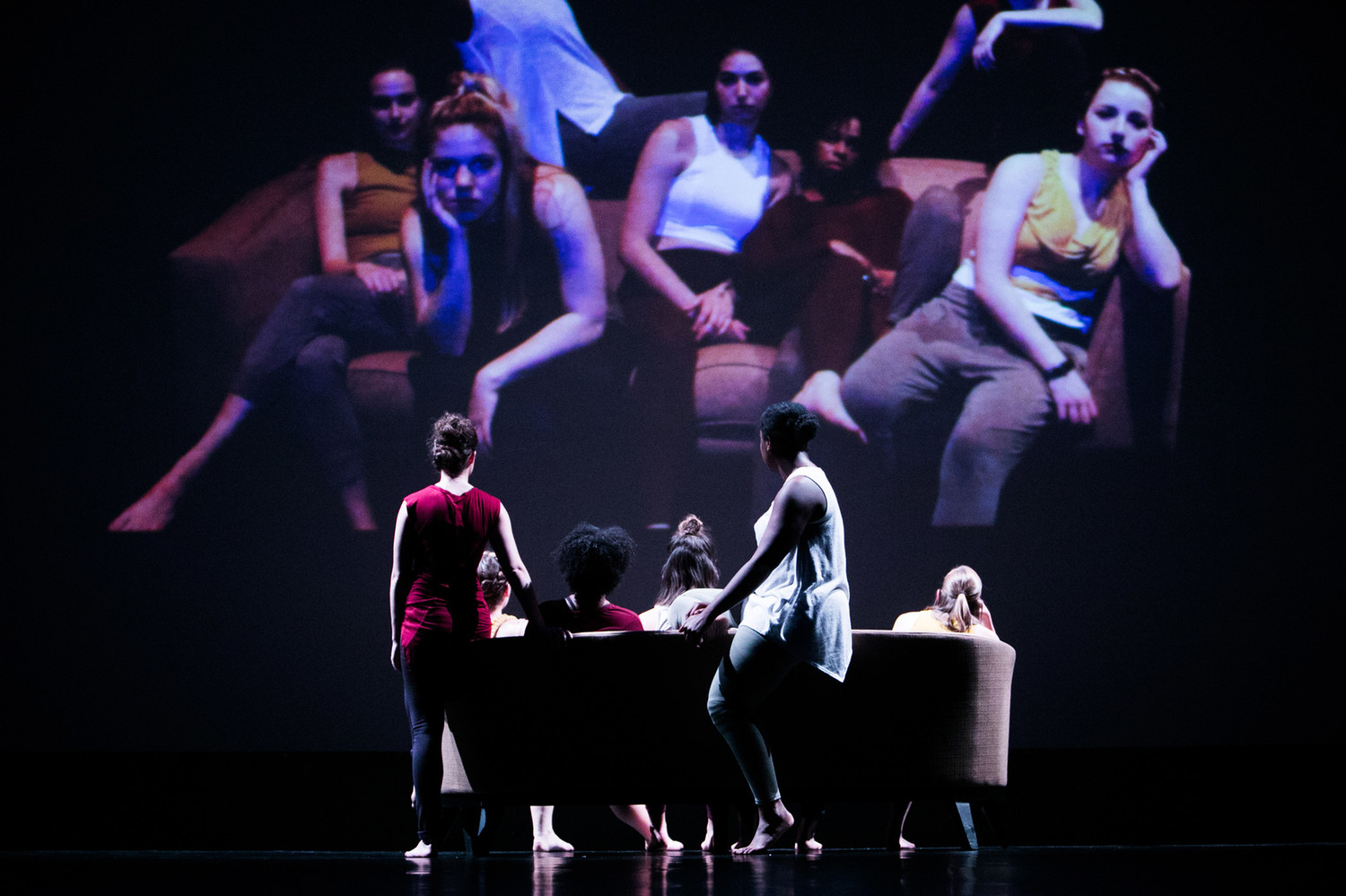 Dancers onstage sitting on couch looking at projection image of themselves